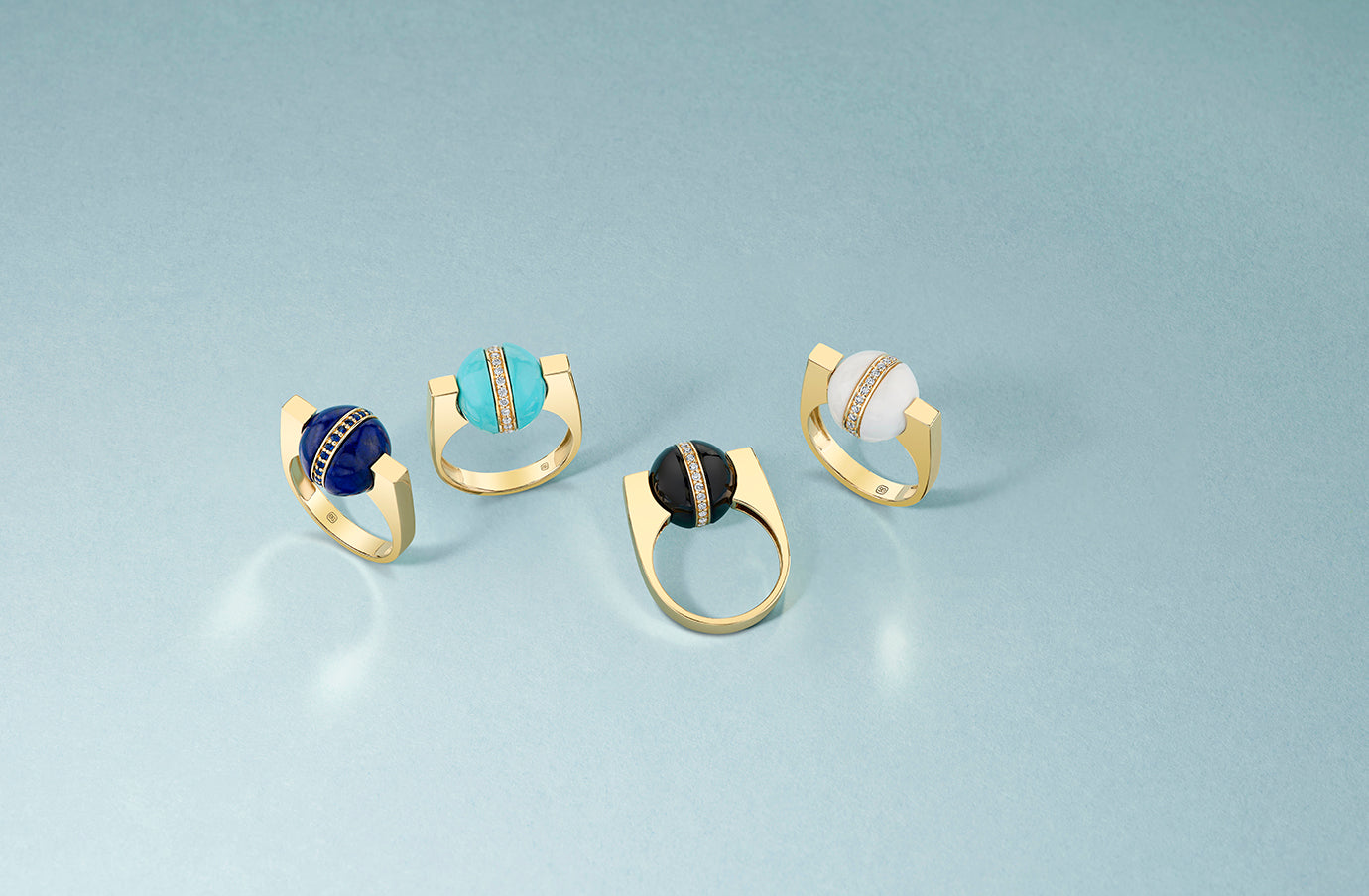 Angled view of four gold rings showcasing colorful gemstone spheres and pavé bands on a blue background